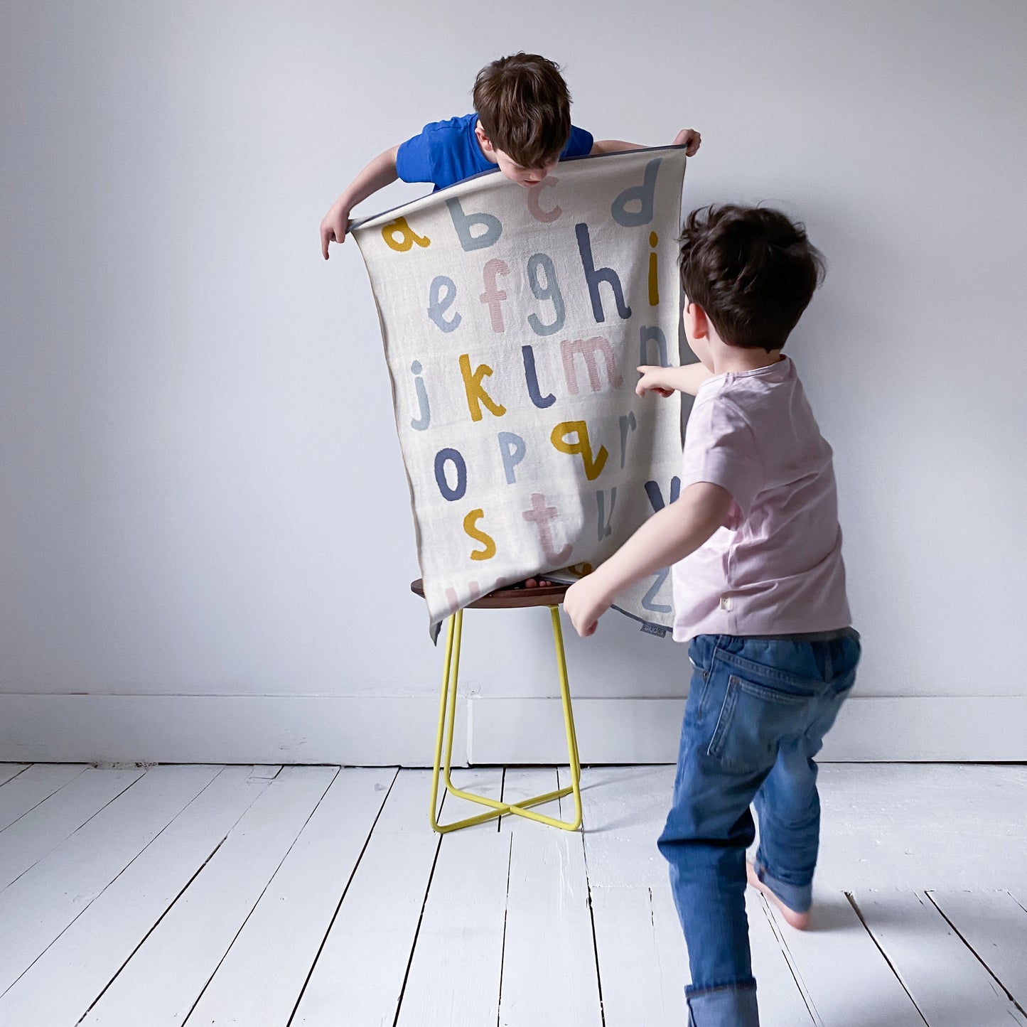 Little boys holding and playing with a Sophie Home Ltd cotton knit alphabet baby pram blanket in cream with mustard, pink and blue letters.