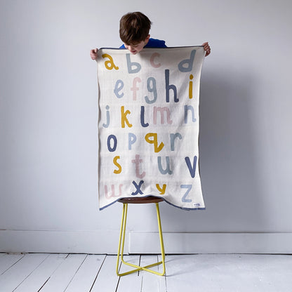 Little boy holding up a Sophie Home Ltd cotton knit alphabet baby blanket in cream with mustard, pink and blue letters.