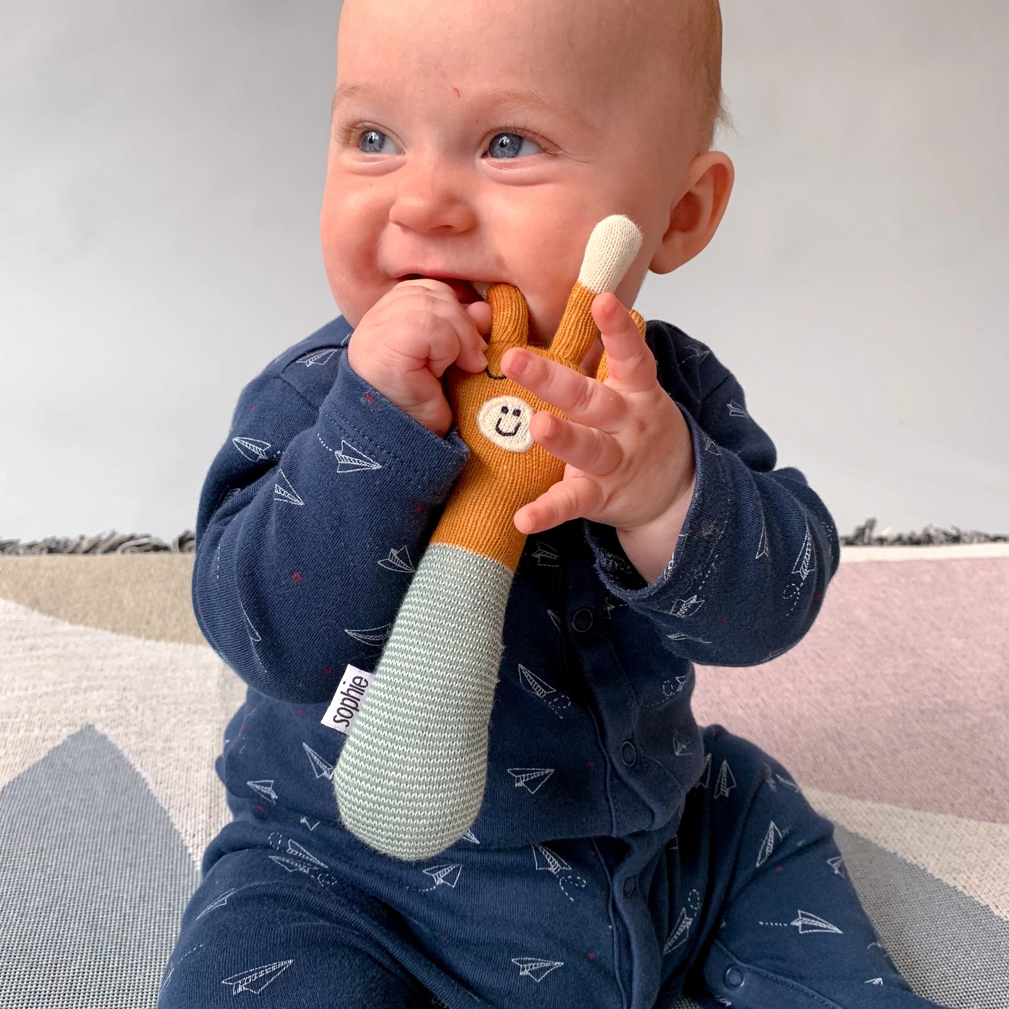 Baby boy holding and chewing Sophie Home Ltd orange and teal cotton knit baby Giraffe Rattle with hand embroidered face.